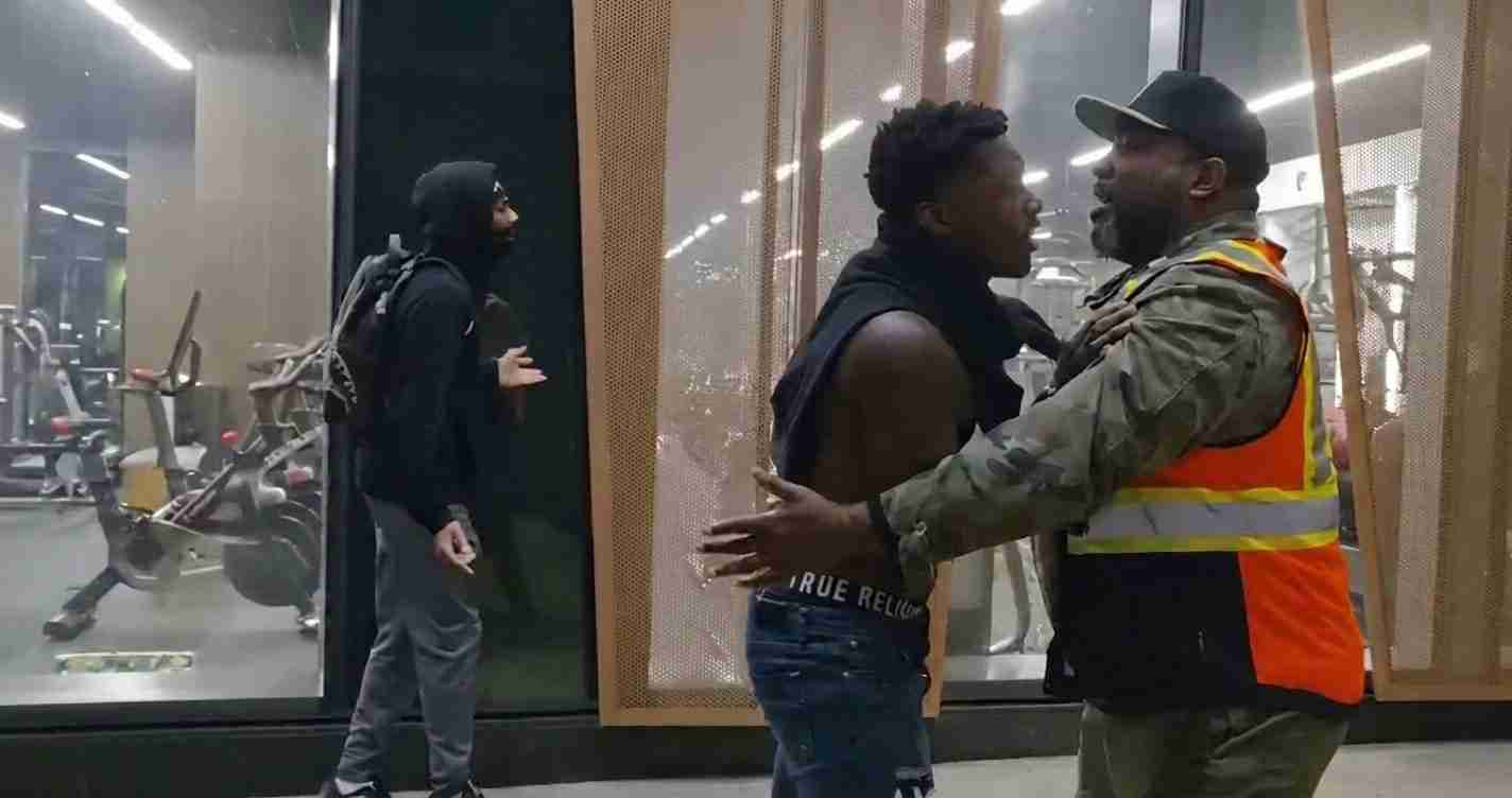 Two men argue in a gym lobby near glass walls, one wearing a bright orange safety vest and cap restraining the other man with his hands on his chest/sides.