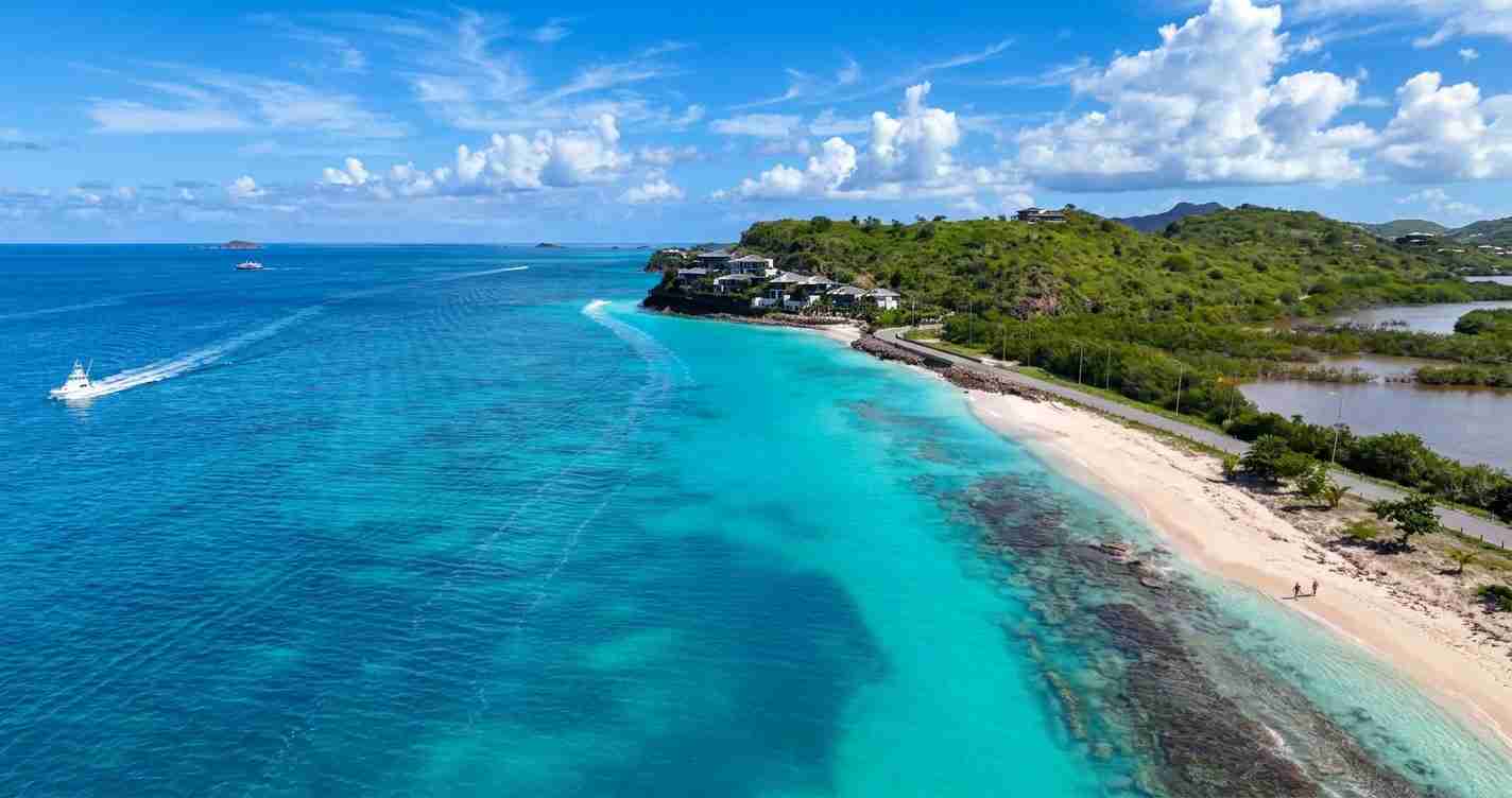 Aerial view of a turquoise-sand beach with green hills and a few buildings along the shore under a blue sky.