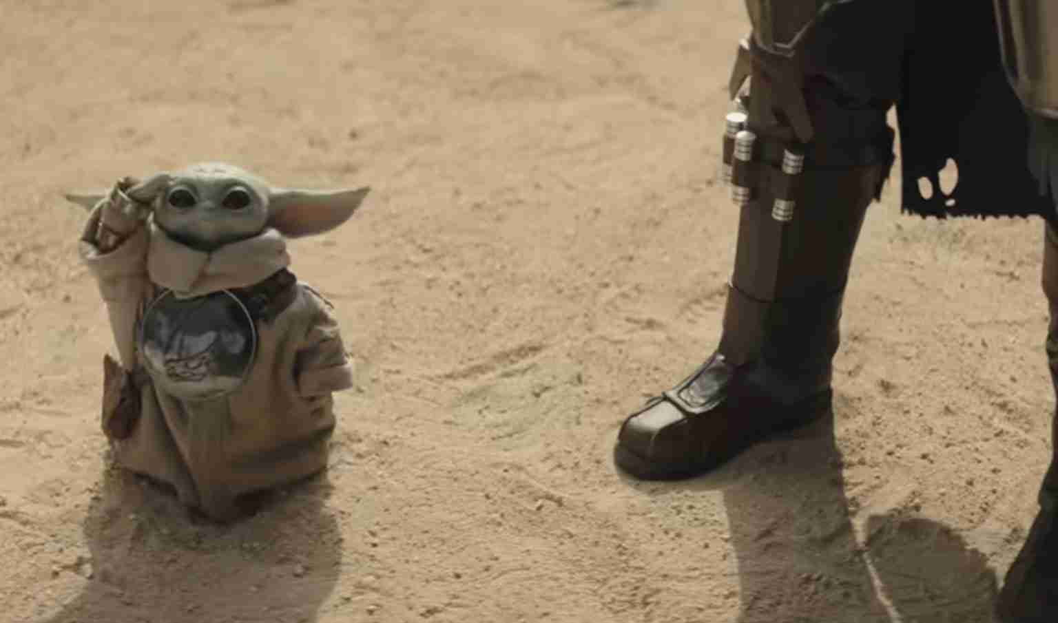 Grogu (Baby Yoda) in a beige robe stands on desert sand, waving beside a armored figure's booted leg.