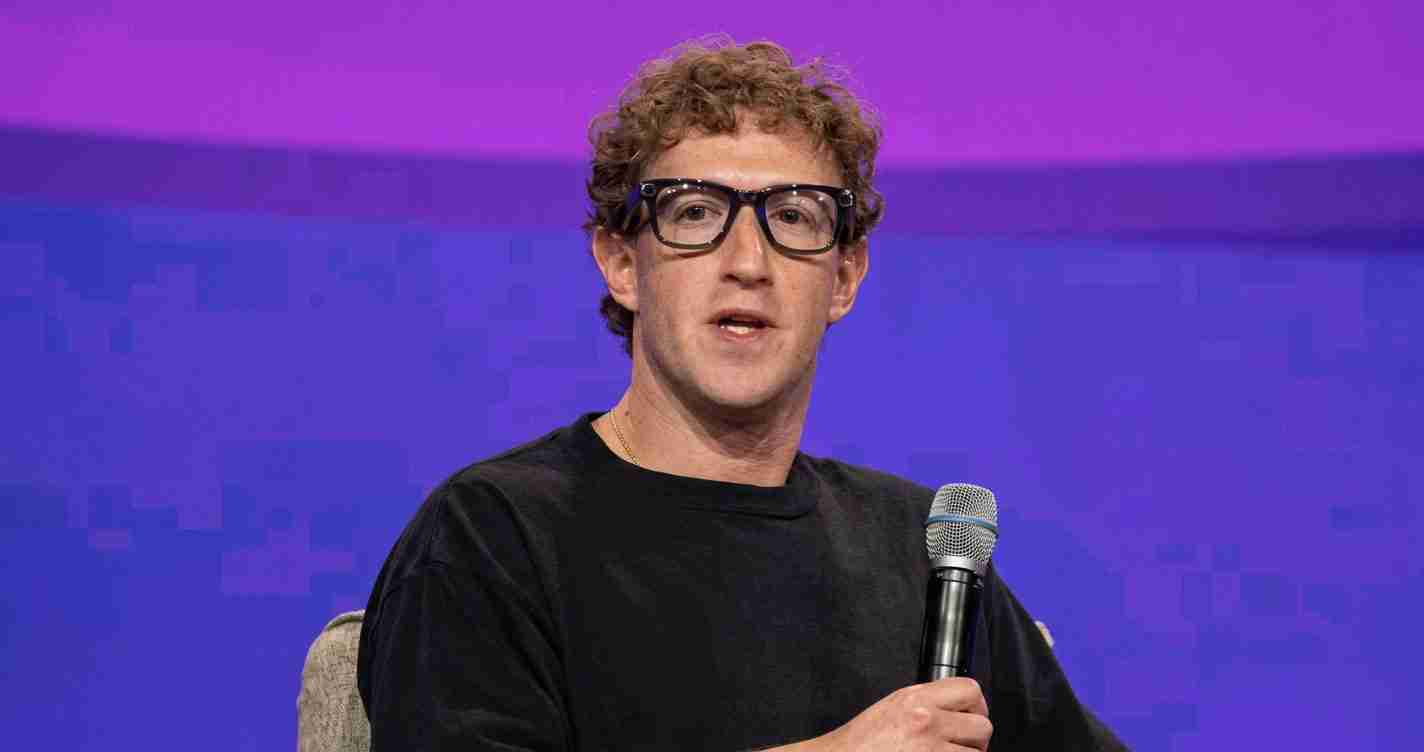 Man in a black shirt with curly hair and thick glasses speaks into a handheld microphone on a purple stage backdrop.