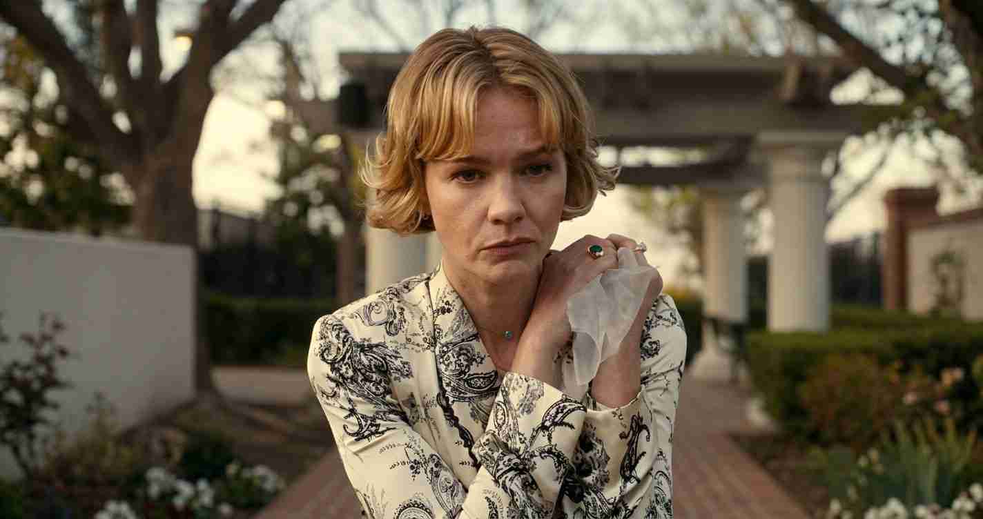 Woman with short blonde hair wearing a patterned blouse stands outdoors, clutching a cloth near her shoulder and looking distressed.
