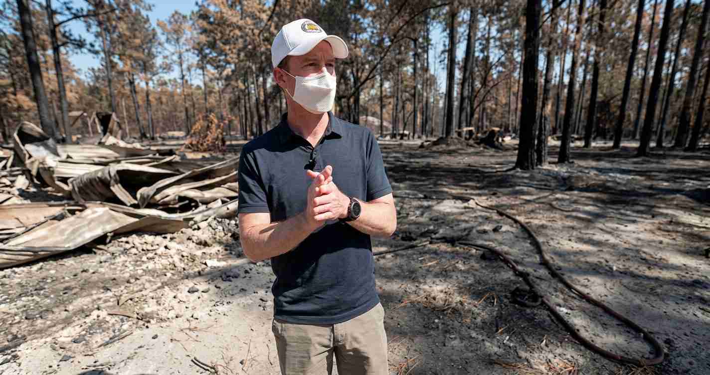 Man in a white cap, navy polo and mask stands in a burned forest with charred trees and debris left behind.