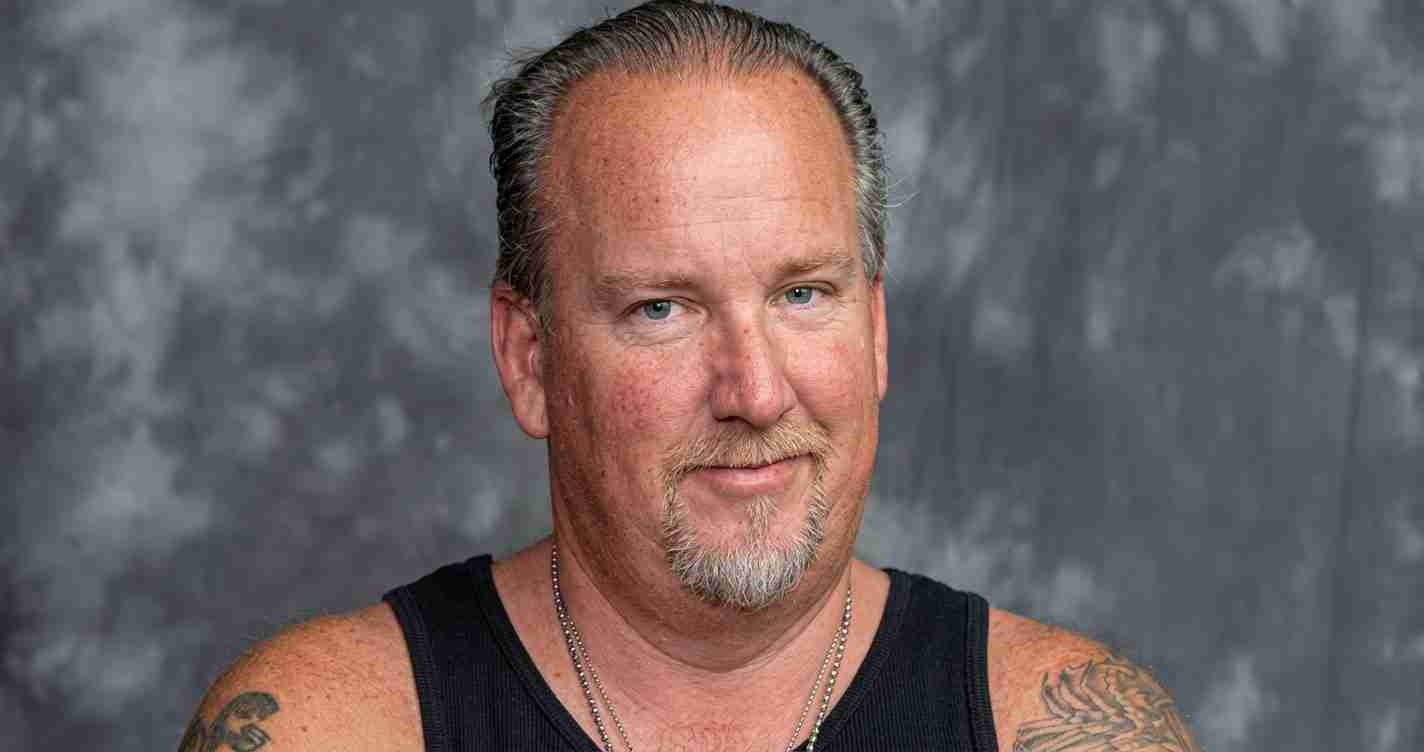 Smiling man with short hair and goatee wearing a black sleeveless shirt against a gray studio backdrop with tattoos on his arms.