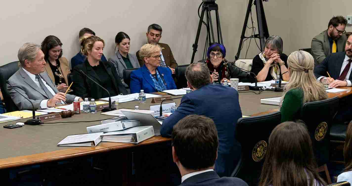 Formal committee meeting around a long conference table with microphones, binders, and attentive attendees.