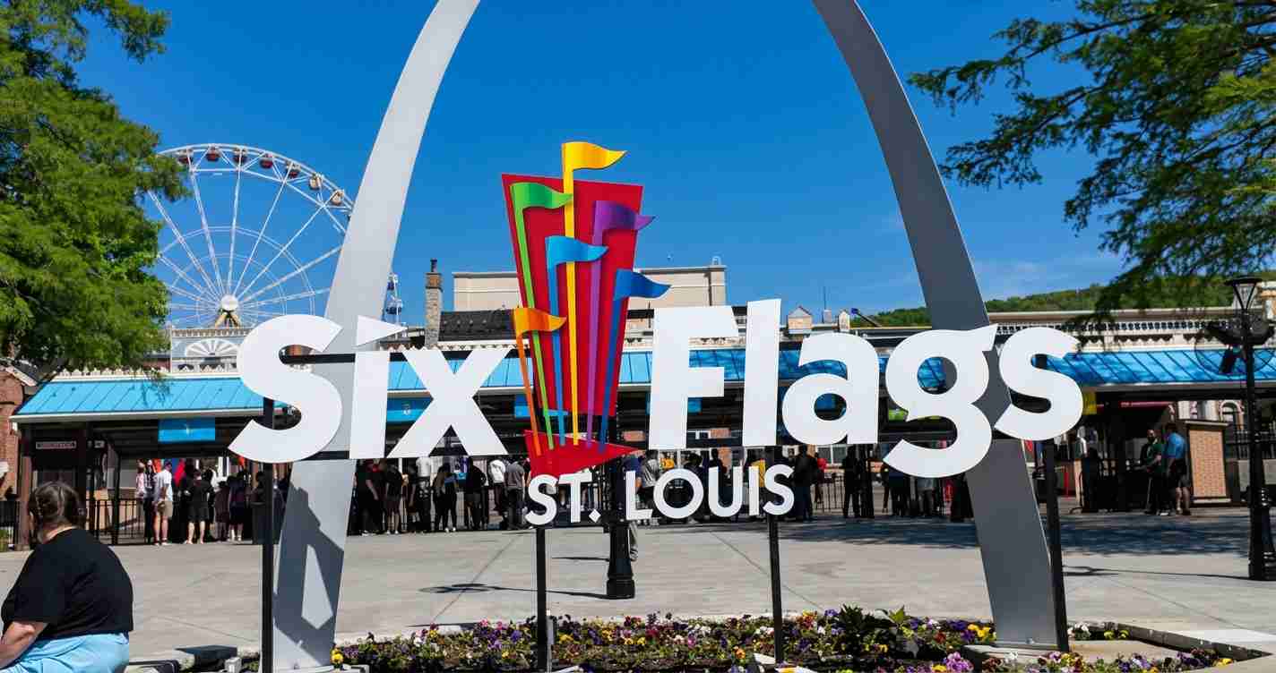 Six Flags St. Louis sign with colorful vertical flags and a backdrop of the entrance and Ferris wheel against a blue sky.