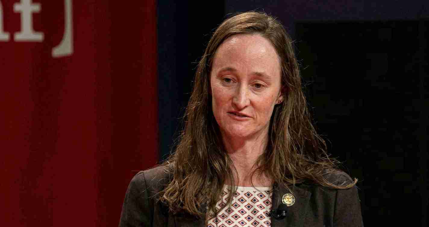 Woman with long brown hair speaking on a stage, wearing a dark blazer and patterned blouse, a lapel pin visible on her left lapel