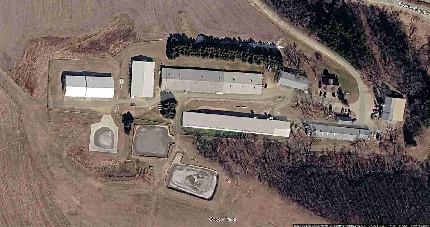 Aerial view of a rural facility with four long white buildings, two large basins, and a parking area among bare fields.