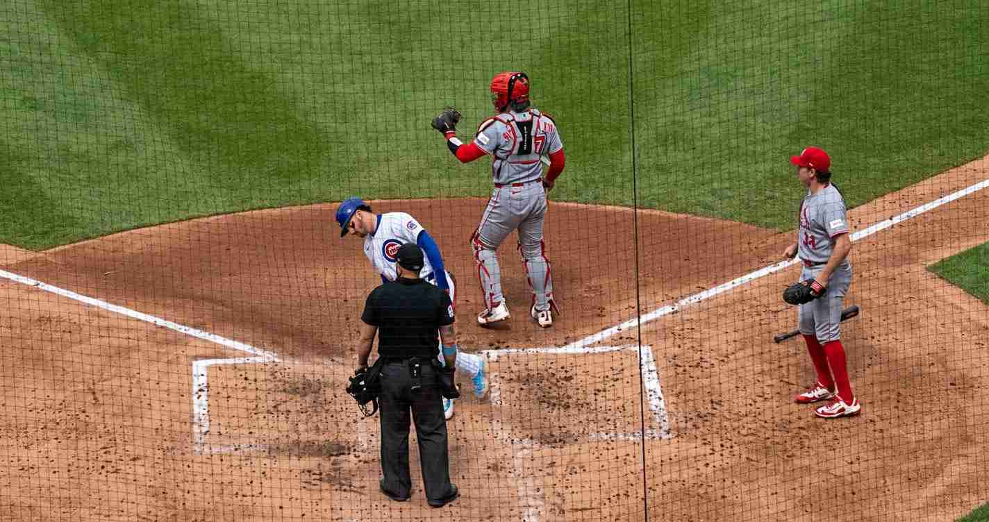 Baseball scene at home plate with umpire, catcher, and baserunner waiting near the plate on a dirt infield, netting in foreground.