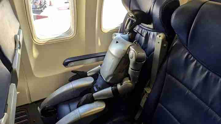 Humanoid robot sitting in an airplane window seat with metal limbs and blue leather seating.