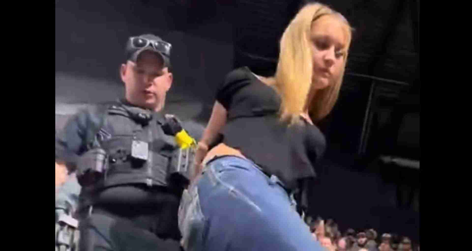Security guard in tactical vest on a stage beside a blonde woman leaning toward the audience at a concert.