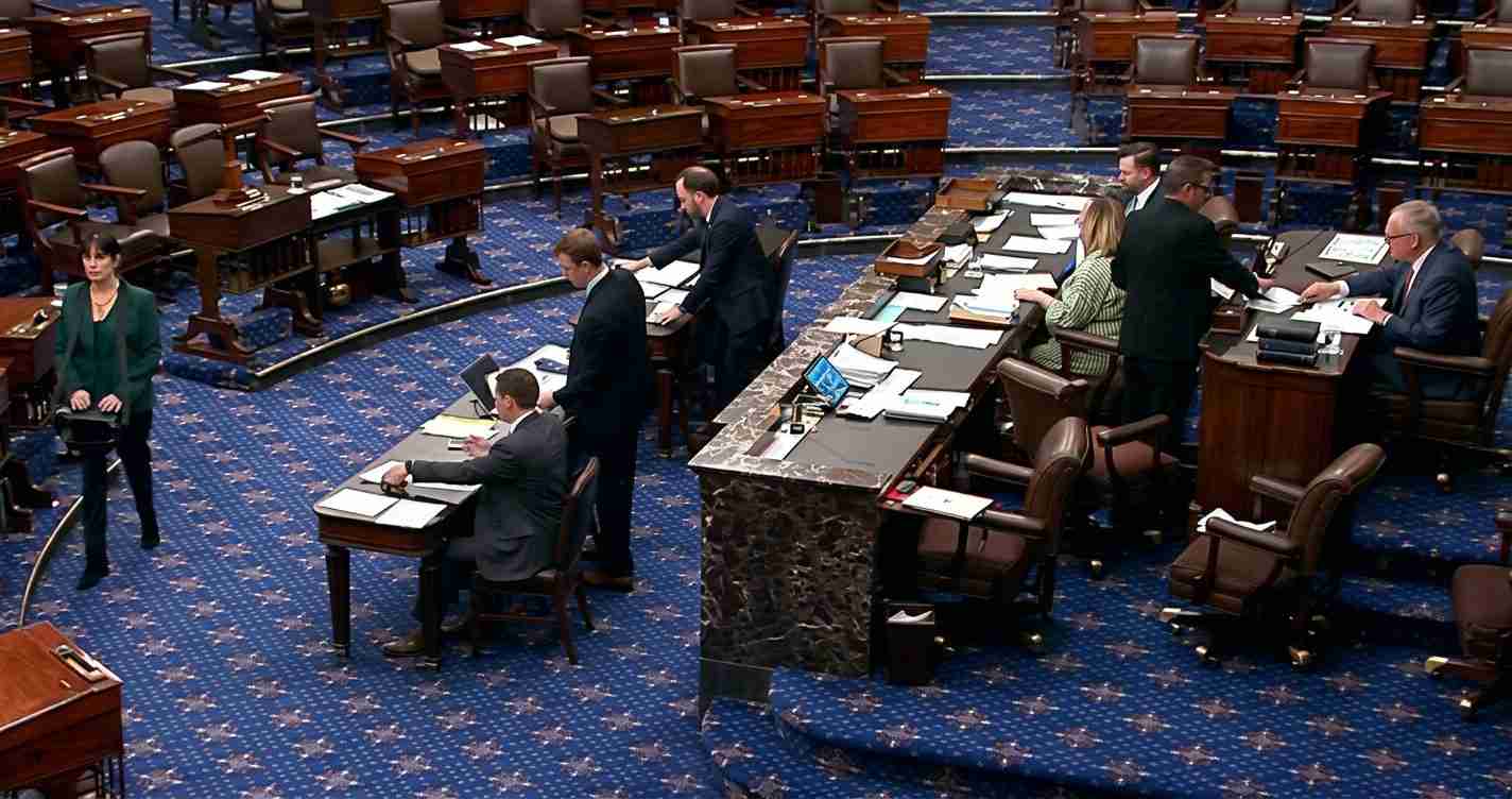 Senate chamber with desks, officials in suits, and a marble podium; blue patterned carpet and arranged seating visible throughout the room.