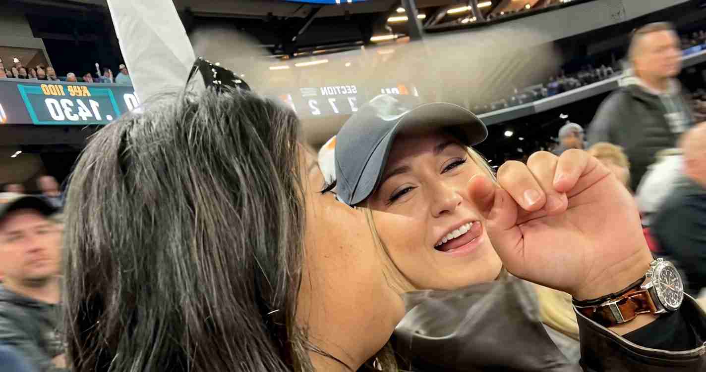 Two women at a crowded stadium share a kiss on the cheek as one leans in, smiling in a gray cap and sunglasses.