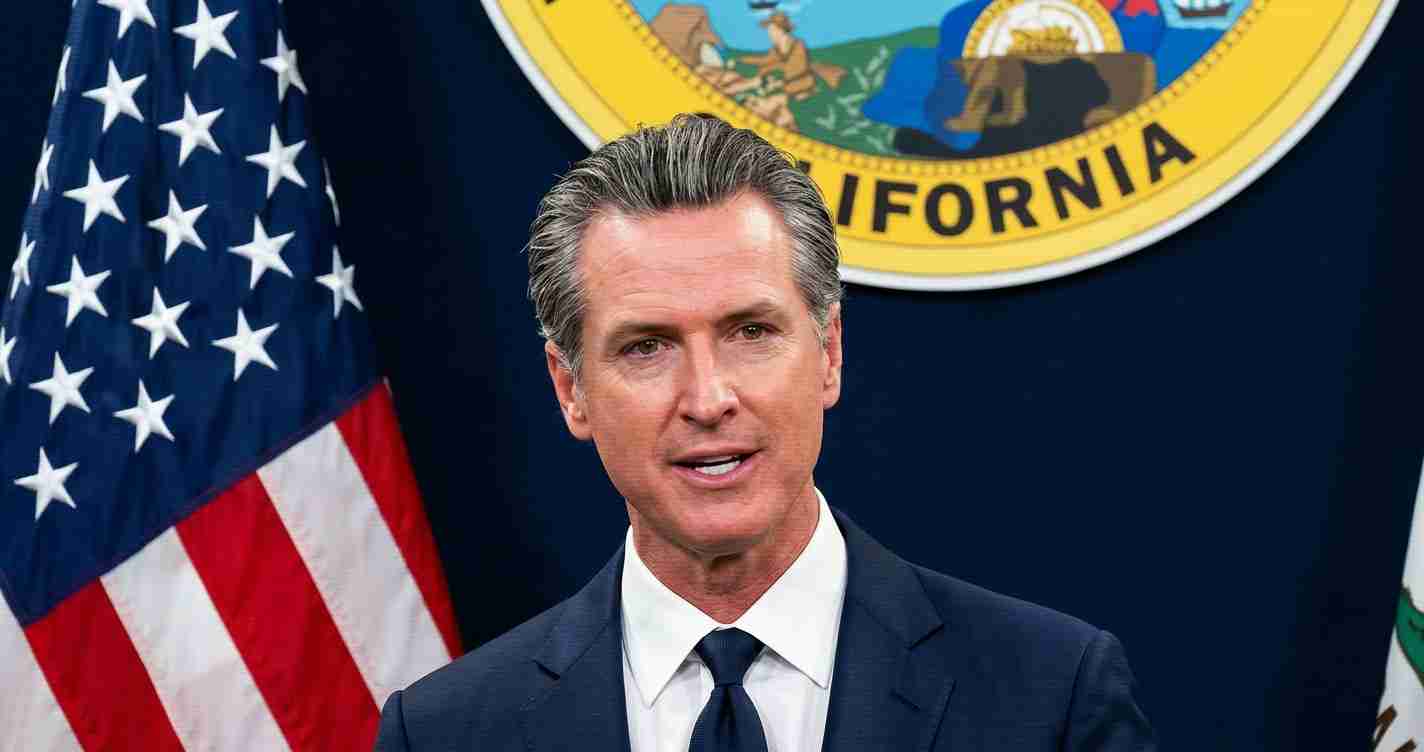 Male politician giving a press briefing, with the California state seal and American flag behind him? behind him visible in background.