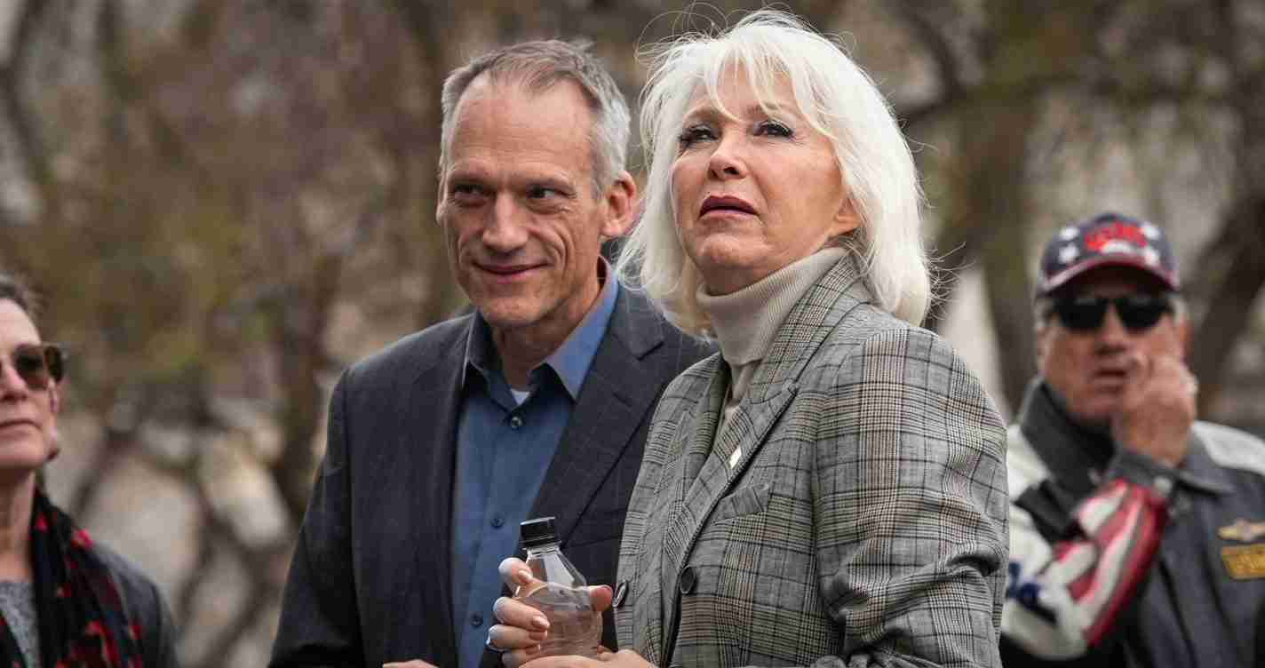 An older woman with white hair wearing a gray plaid blazer, holding a water bottle, standing beside a man in a blue shirt outdoors.