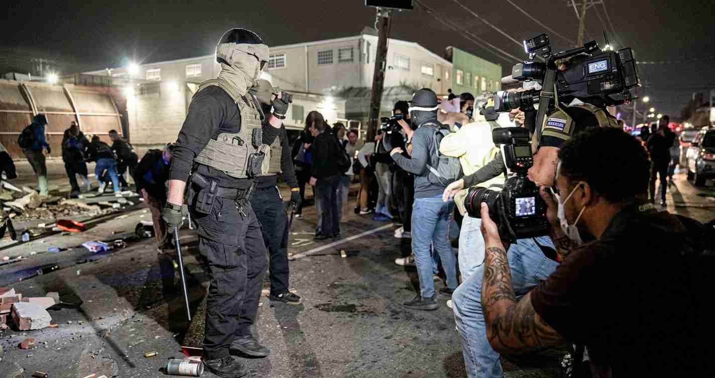 Night scene of a heavily armed officer in tactical gear facing a line of photographers and reporters on a debris-strewn street.