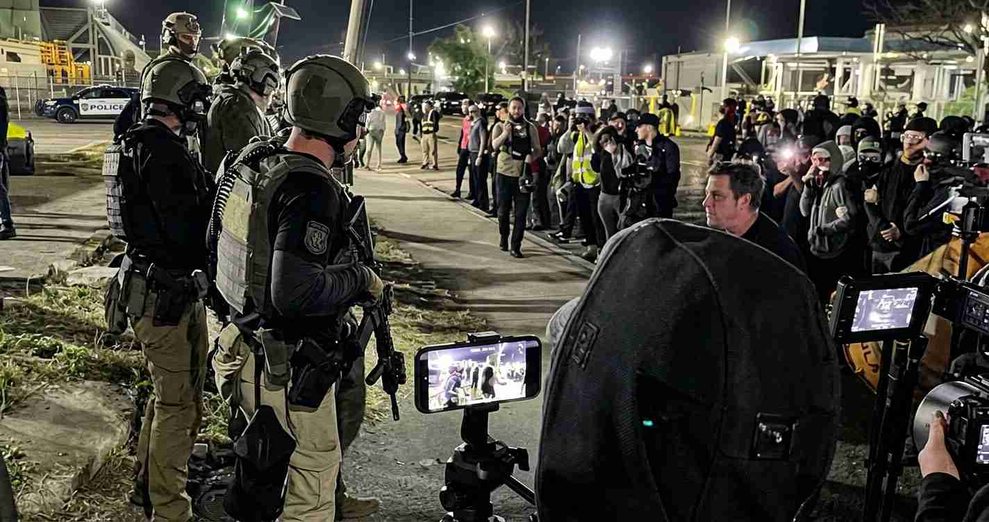 Line of heavily armed officers in tactical gear facing a crowd of reporters and onlookers at night, cameras flashing in the background.