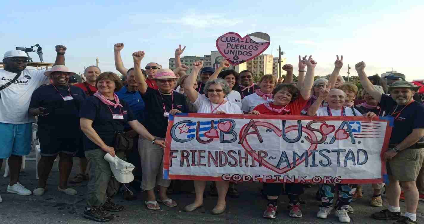 Group of diverse people at a sunny rally holding a banner reading Cuba-USA Friendship/Amistad with hearts and flags in the corners.