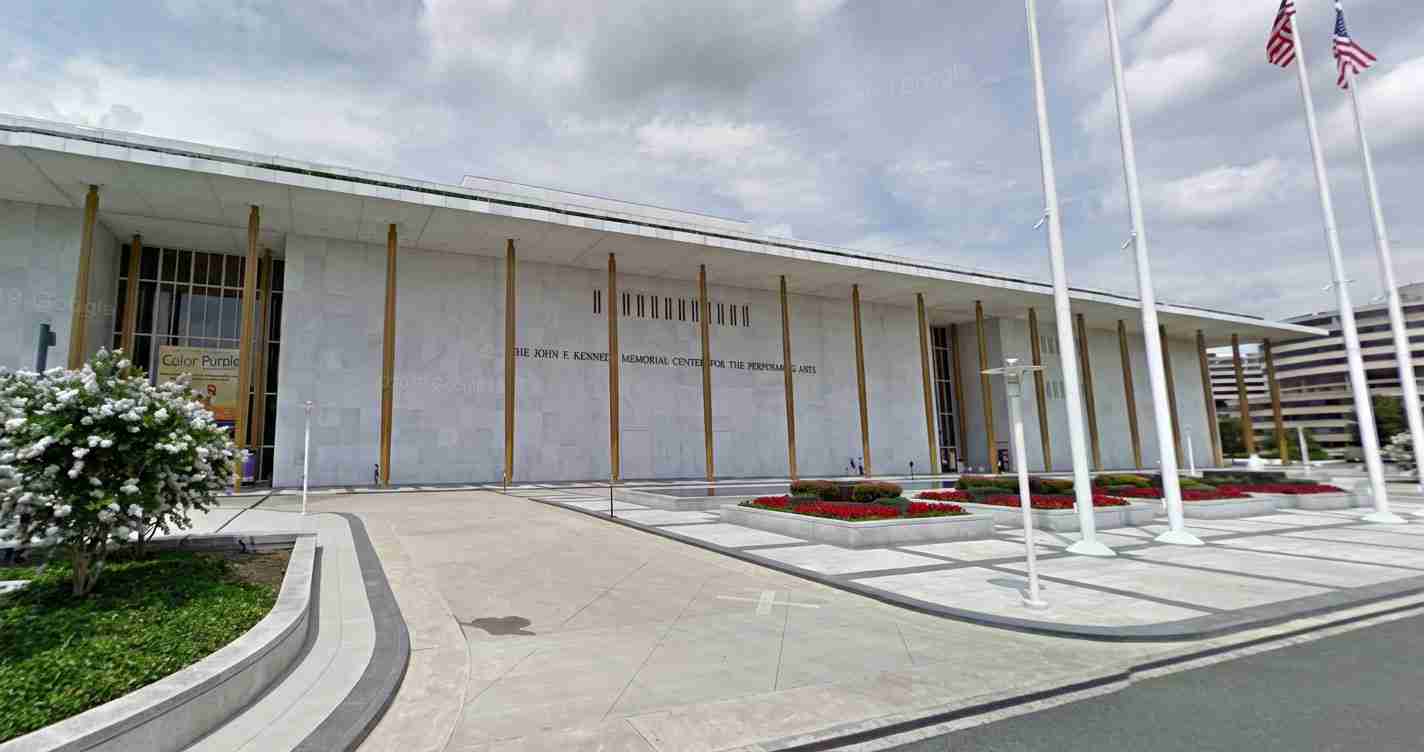 Exterior view of a modern memorial center with tall gold columns, glass doors, and flower beds along a wide walkway under a cloudy sky and flags nearby.