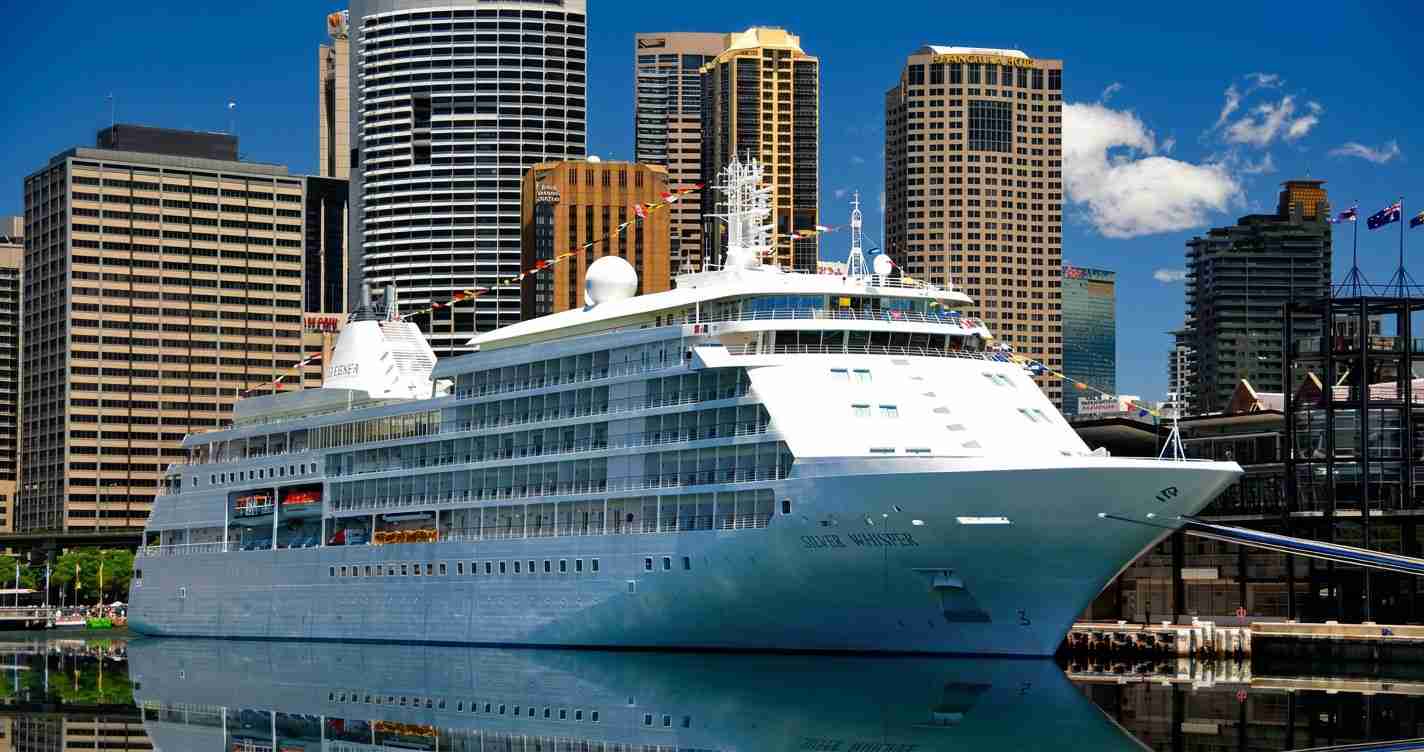 A large white cruise ship docked at a harbor with a city skyline in the background.