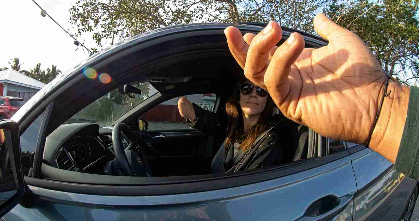 Driver wearing sunglasses sits in a blue car with the window down, reaching a hand toward the camera as sunlight creates lens flare.