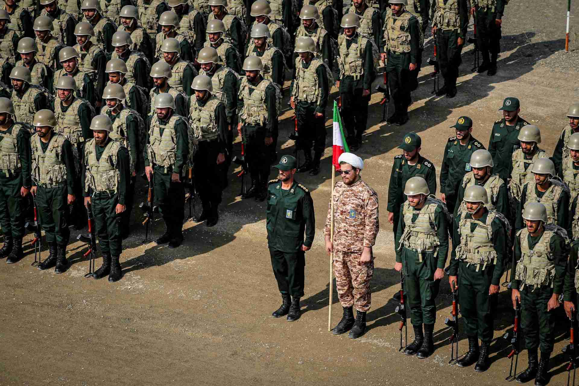 Rows of soldiers in green uniforms and helmets stand at attention on a dusty field; a flag bearer in camouflage leads a line of troops nearby.