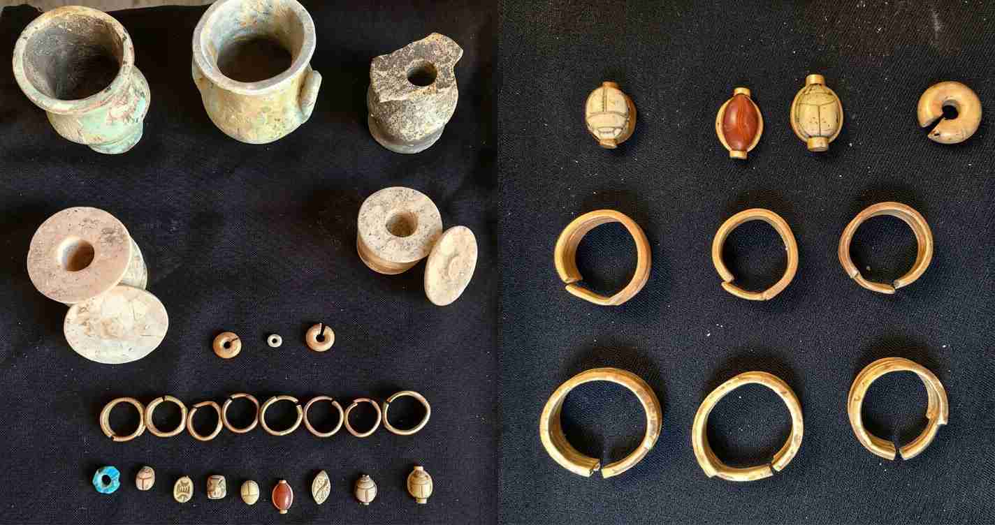 Collection of ancient jewelry and vessels laid out on dark fabric: bowls, rings, and beads visible on left and right sections.