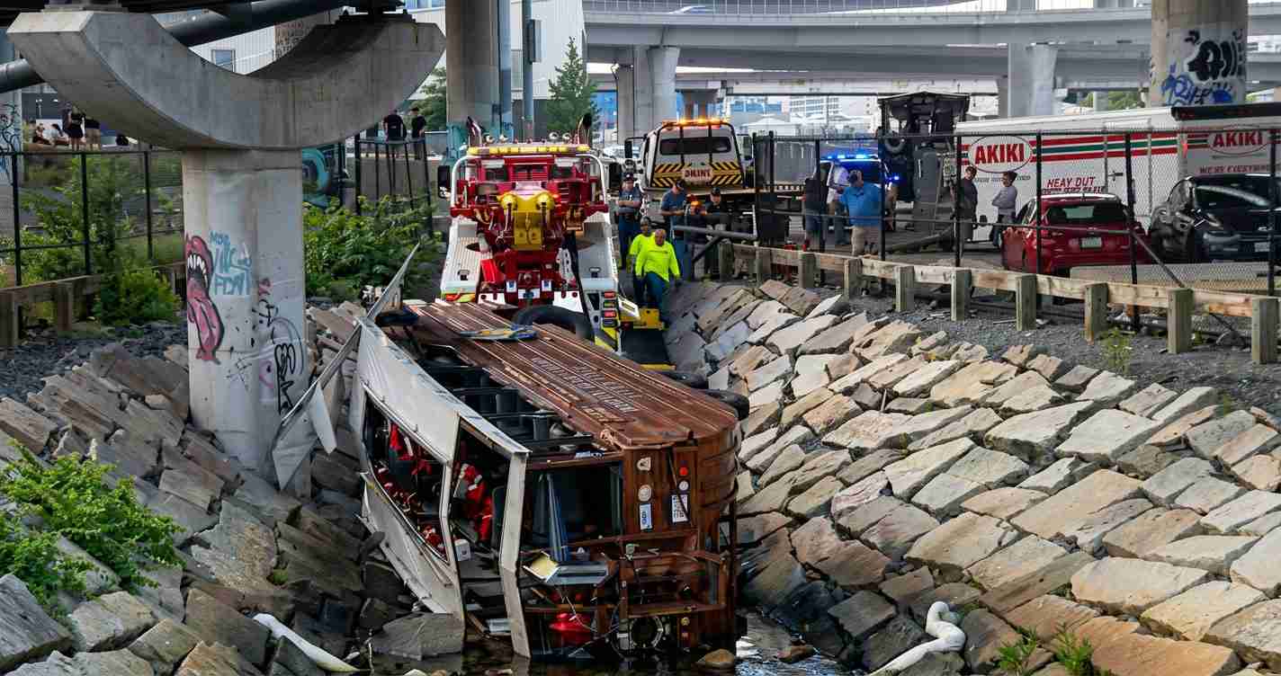 Overturned bus in a rocky canal beside a highway, with emergency crews and rescue vehicles on scene.