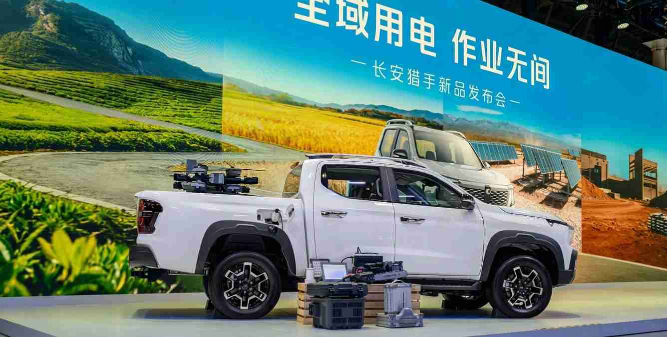 White pickup truck on a showroom stage with equipment mounted in the bed, set against a large blue exhibition banner with Chinese text and a landscape backdrop.