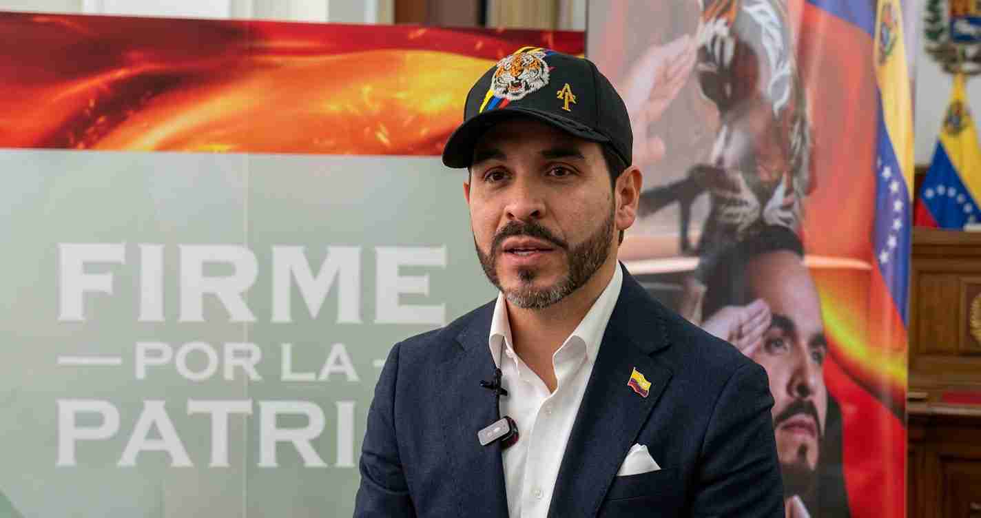 Man in a navy blazer speaking in an interview, wearing a black cap with a tiger emblem and a Venezuelan flag pin, with a banner reading 'FIRME POR LA PATRIA' in the background.