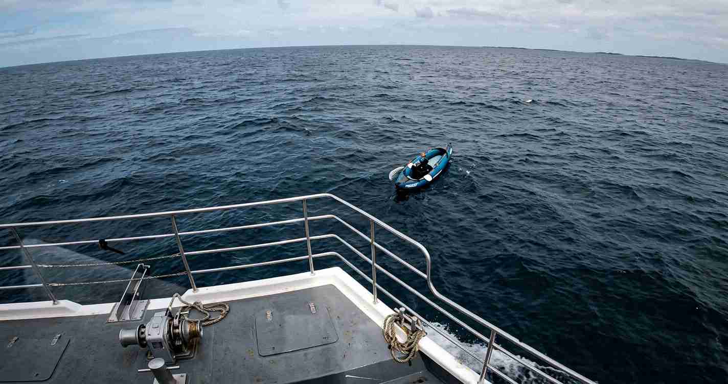 View from a larger ship's deck showing metal railings and a small blue inflatable dinghy drifting in open ocean nearby with a rope and winch on the deck foreground side.