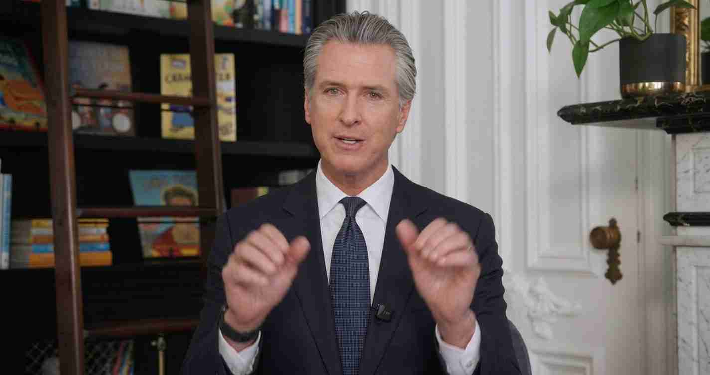 Man in a dark suit and tie speaks to camera, hands mid-gesture, in a room with bookshelves behind him.