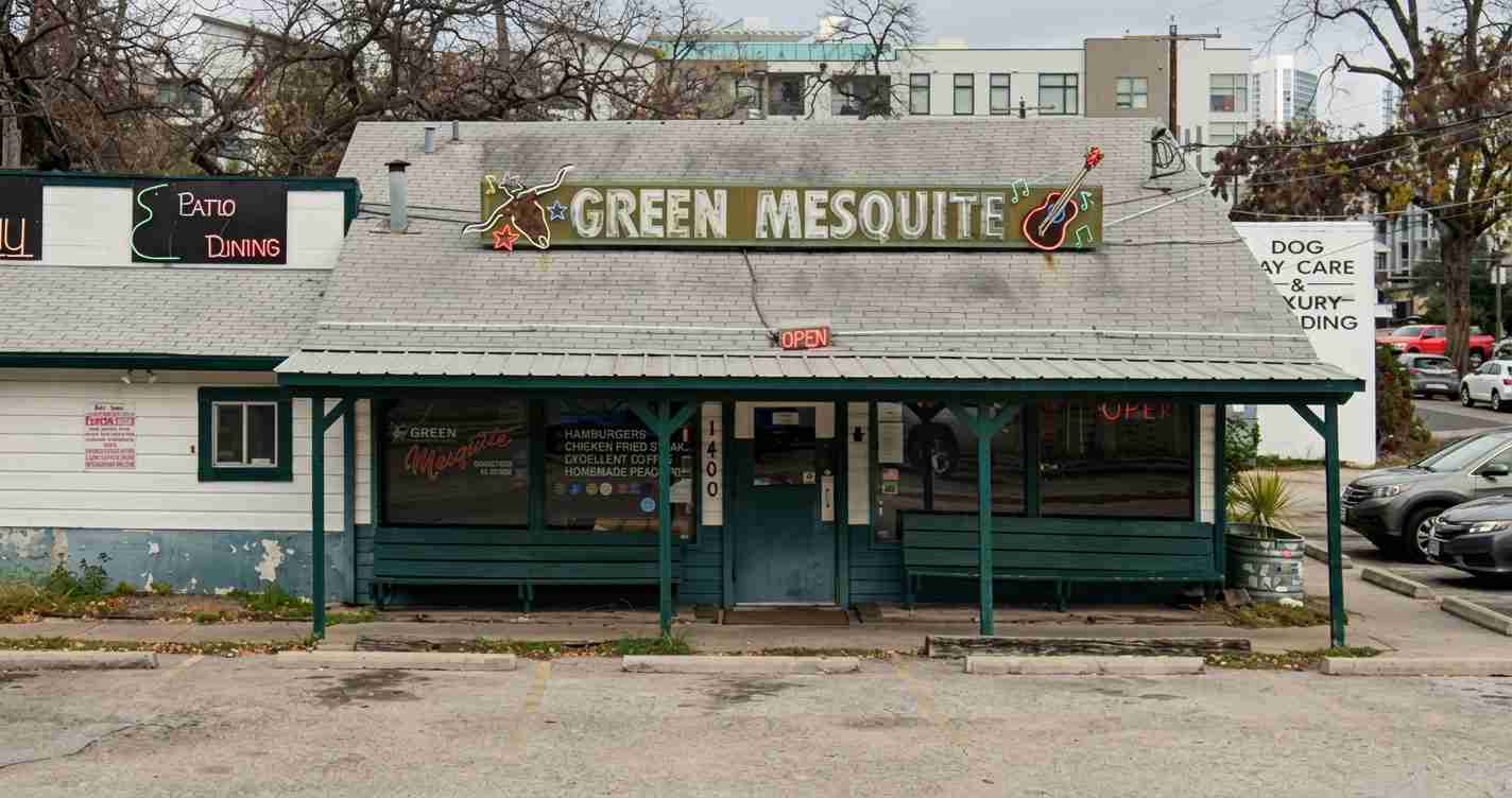 Front of a small, teal-painted restaurant with a large, neon-style 'GREEN MESQUITE' sign and a hanging 'OPEN'; patio seating visible under the awning.