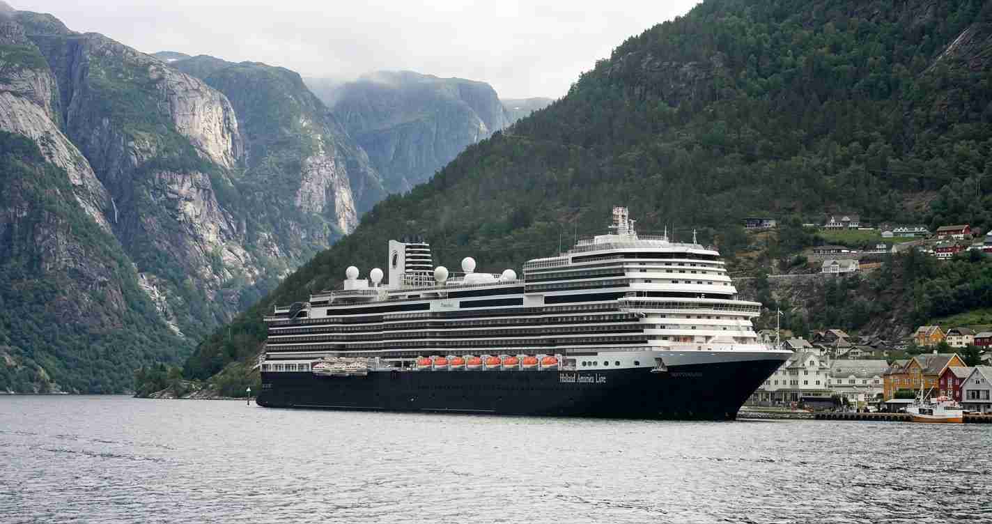 Large cruise ship anchored in a fjord, with steep forested cliffs and a small town along the shoreline.