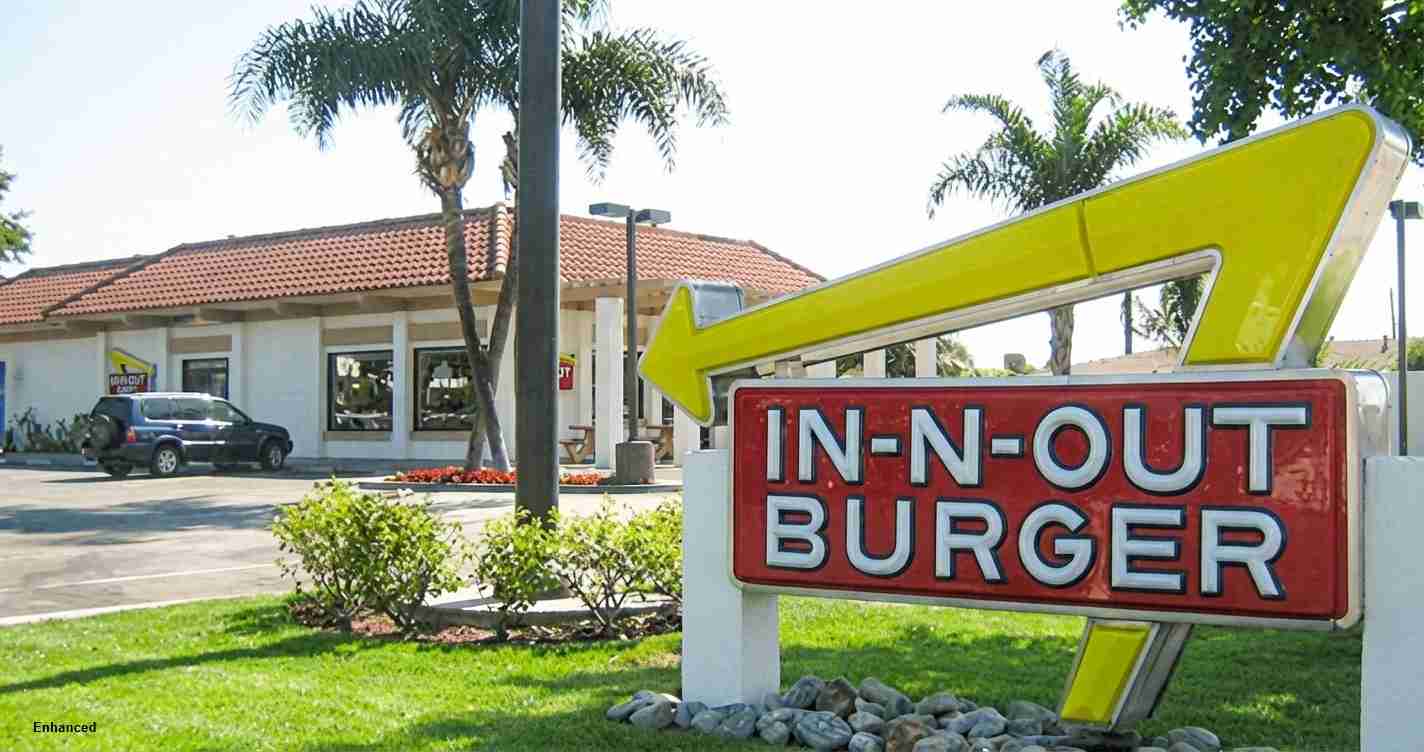 In-N-Out Burger sign with a large yellow arrow in front of a sunny strip-mall building and palm trees.