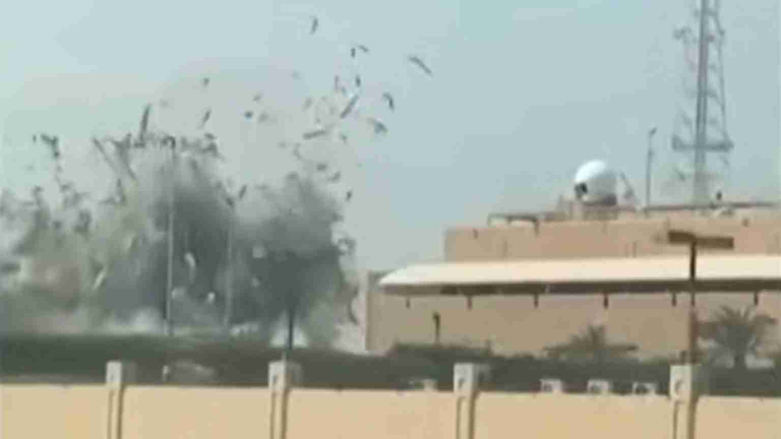 Explosion and smoke near a beige military-style building, palm trees, and a fence in the foreground; birds are scattering in the air.