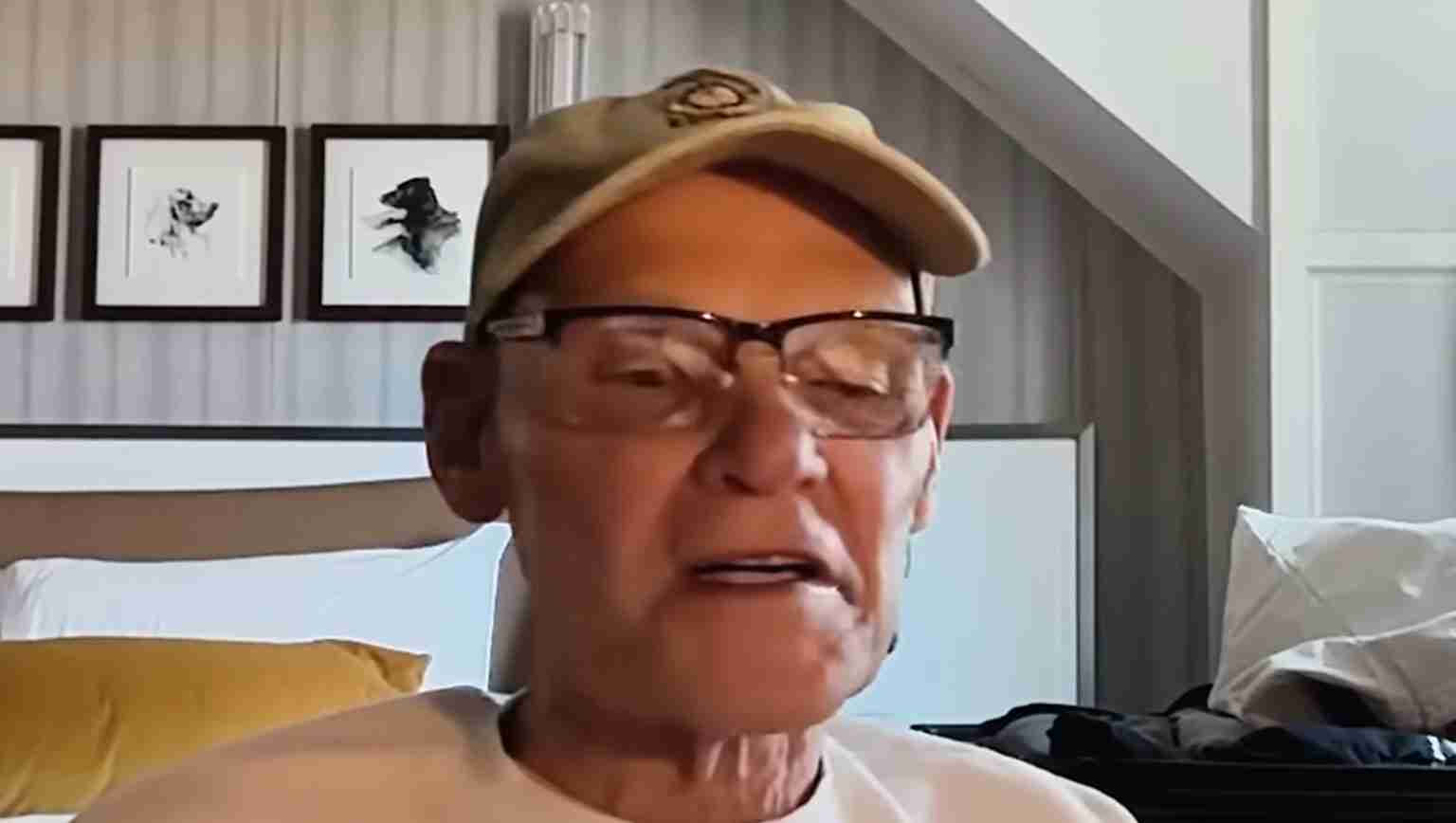 Older man wearing a beige baseball cap and glasses speaks during a video call in a bedroom.