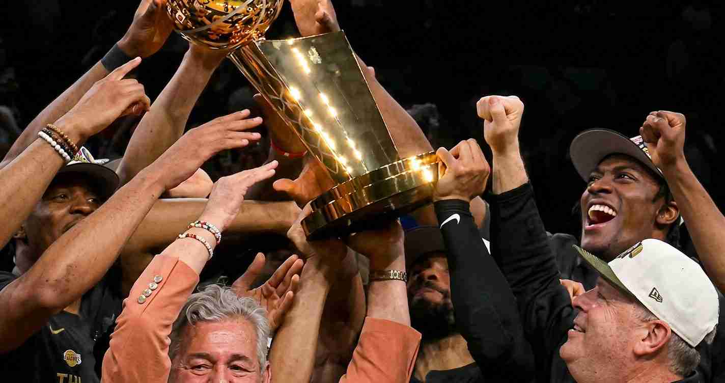 A group of athletes lifts a large golden trophy in celebration, hands reaching up together.