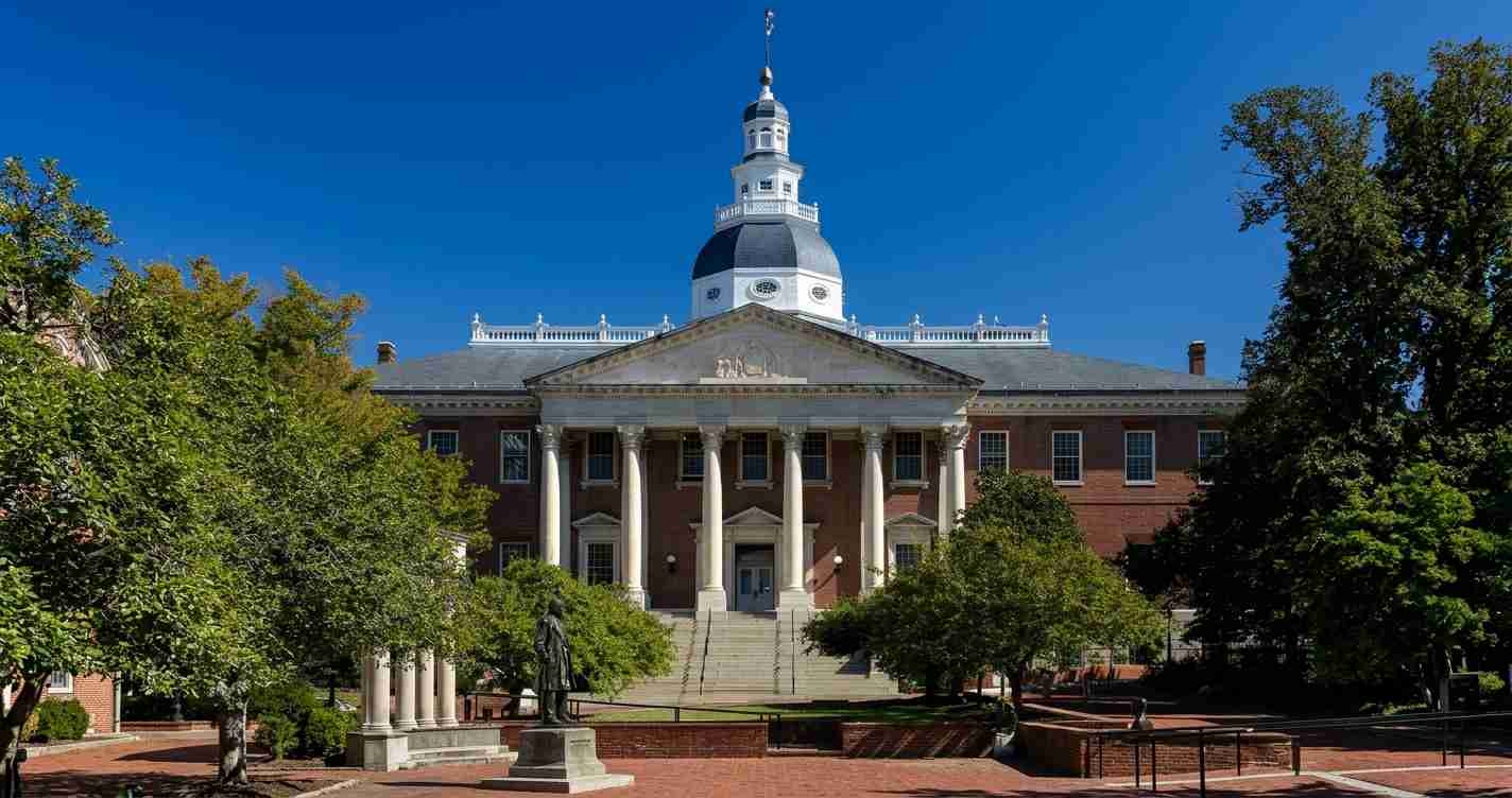 Front view of a brick university building with tall white columns, a pediment, and a central domed tower under a blue sky, surrounded by trees and steps.