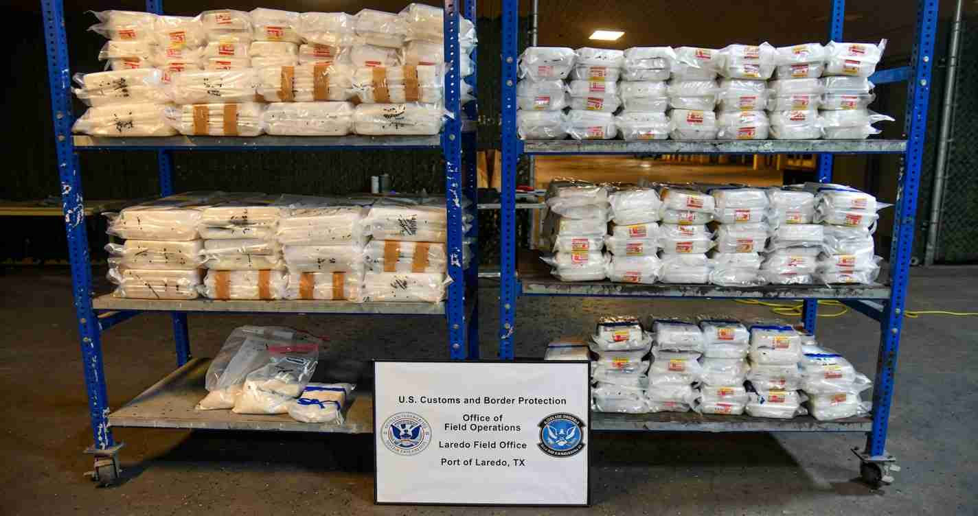 Stacks of white bags on blue metal shelves in a warehouse, with a sign for U.S. Customs and Border Protection (Laredo Field Office).