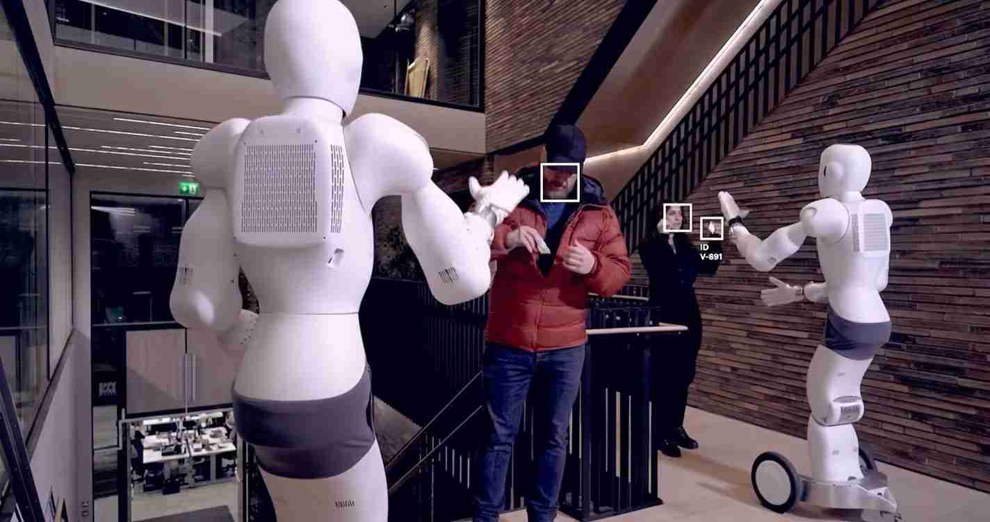 Two white humanoid robots face a man in a red jacket in a modern interior, appearing to interact or demonstrate a gesture in a hallway.