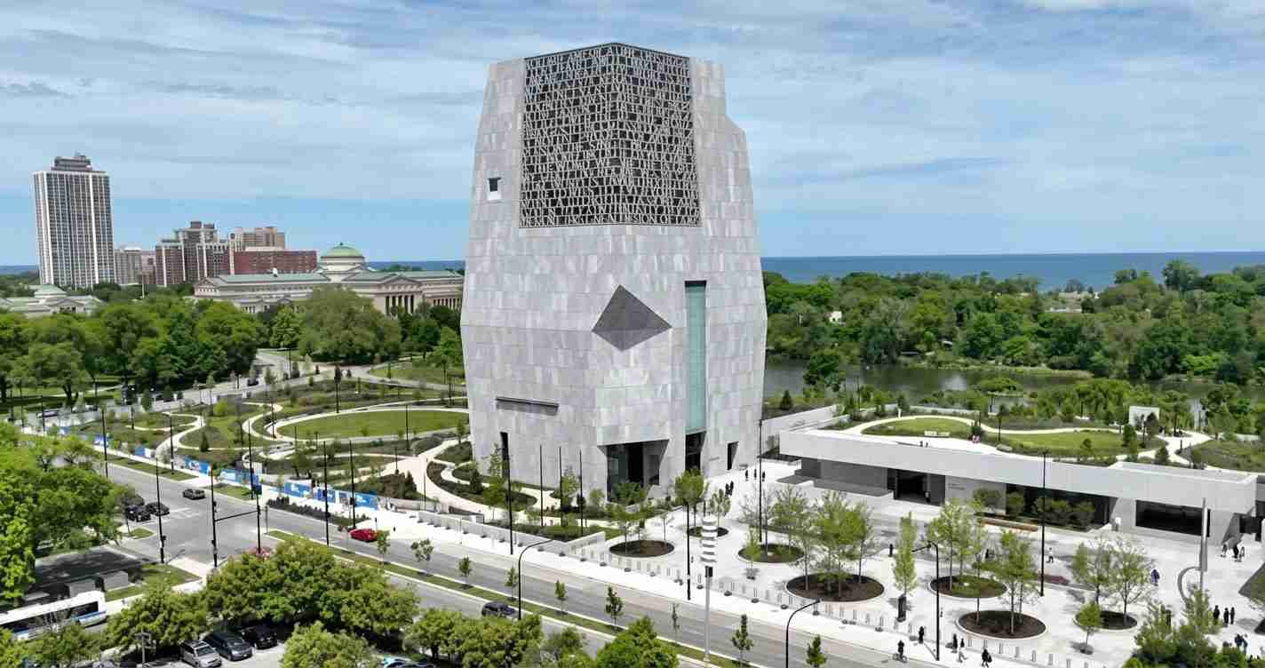 A tall gray modern tower with a lattice upper section, set in a landscaped urban park with roads and trees nearby and water in the distance.