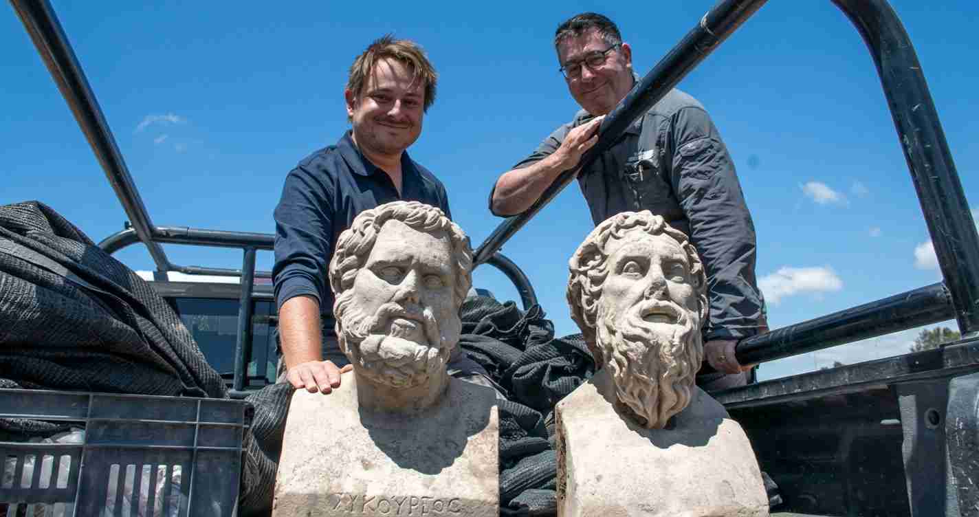 Two workers in a pickup truck bed beside two marble busts of ancient Greek philosophers during a move.