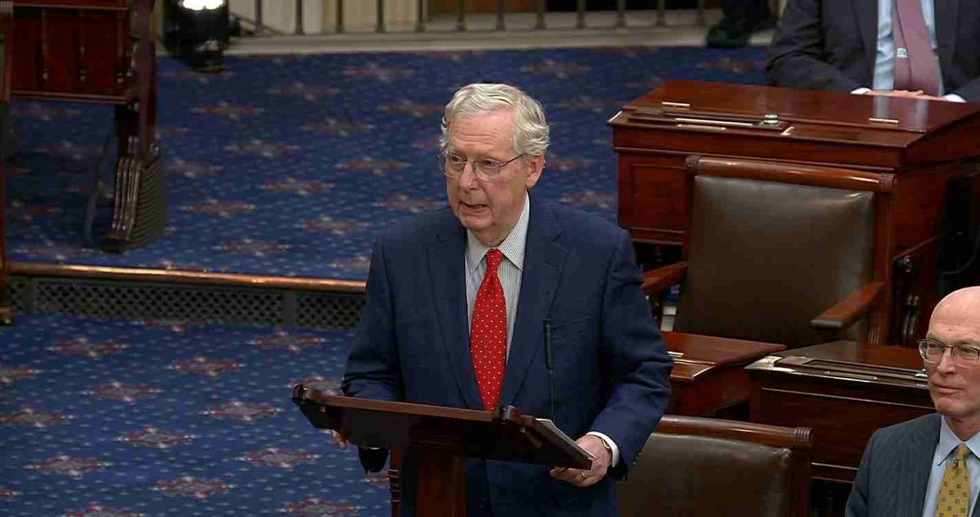 Older man in a navy suit and red tie speaks at a wooden lectern in a legislative chamber on a blue carpeted floor.