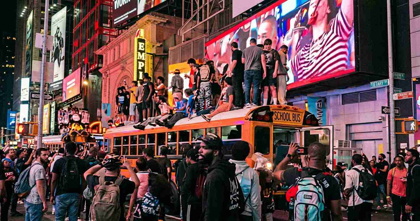 Crowd of people on and around a yellow school bus in Times Square, with bright neon billboards behind them, some taking photos.