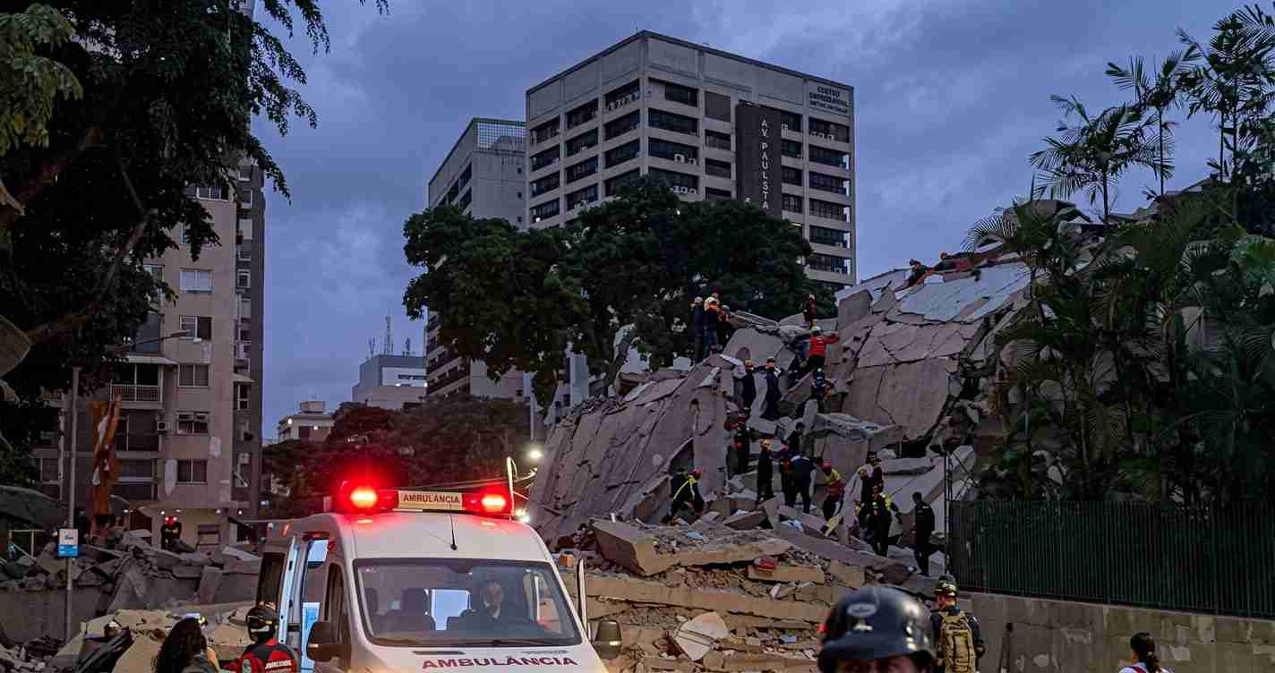 Emergency responders search through a collapsed building with a rubble pile and an ambulance in the foreground.