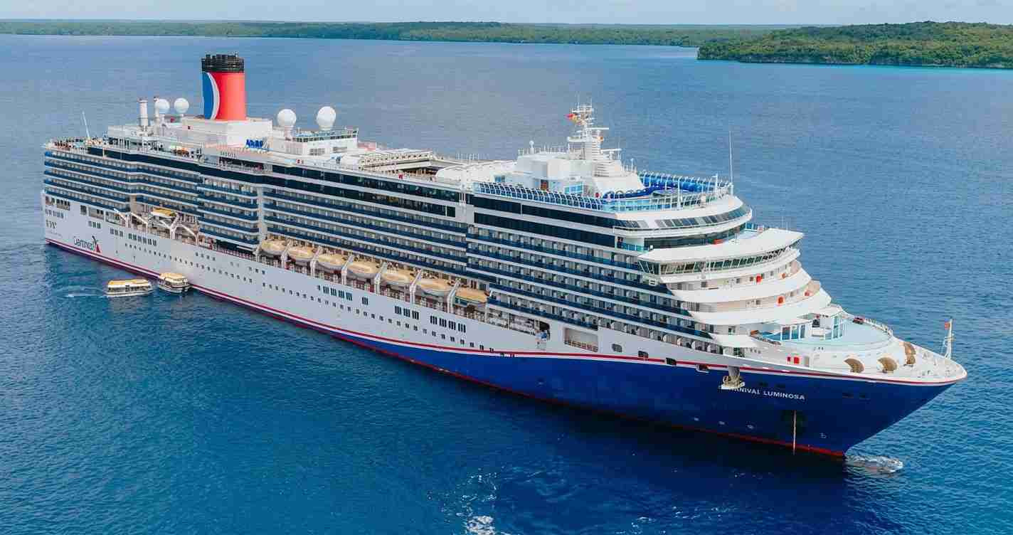 Large white and blue cruise ship with many decks sailing in calm blue water, name Carnival Luminosa visible on the bow.