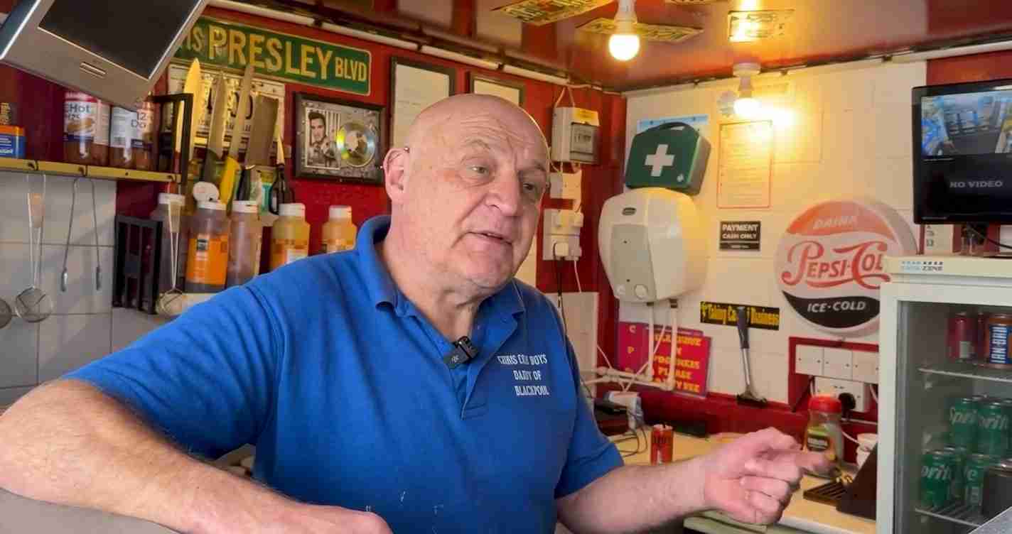 Older man in a blue polo behind a counter in a retro, cluttered shop or bar with neon signs and a Pepsi-Cola sign on the wall.