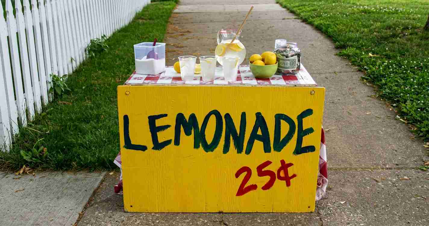 Yellow lemonade stand on a sidewalk with cups, lemons, and a jar of money; 'LEMONADE 25¢' painted on the front.