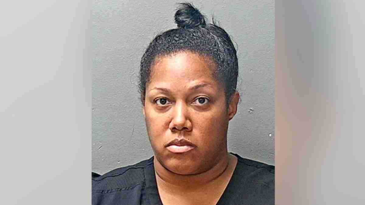 Mugshot of a Black woman with dark hair in a top bun, wearing a black top, facing the camera against a gray background.