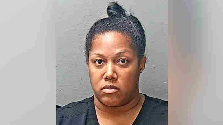 Mugshot of a Black woman with dark hair in a top bun, wearing a black top, facing the camera against a gray background.